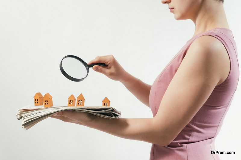 The woman with magnifier holds a newspaper with paper houses in front of yourself. Concept of real estate, housing development.