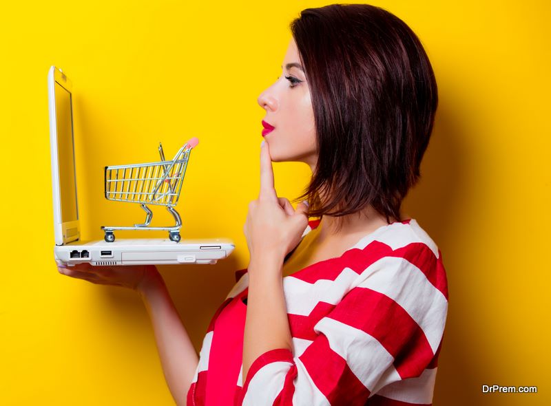 portrait of the beautiful young woman with cart for shopping and laptop on the yellow background